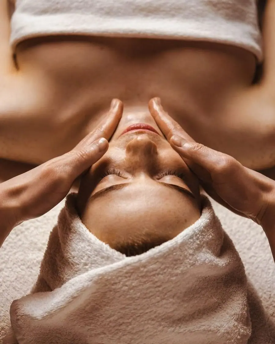 Overhead view of a client lying back with her eyes closed and her hair wrapped in a white towel, a therapist’s hands working a facial massage across her cheeks and temples